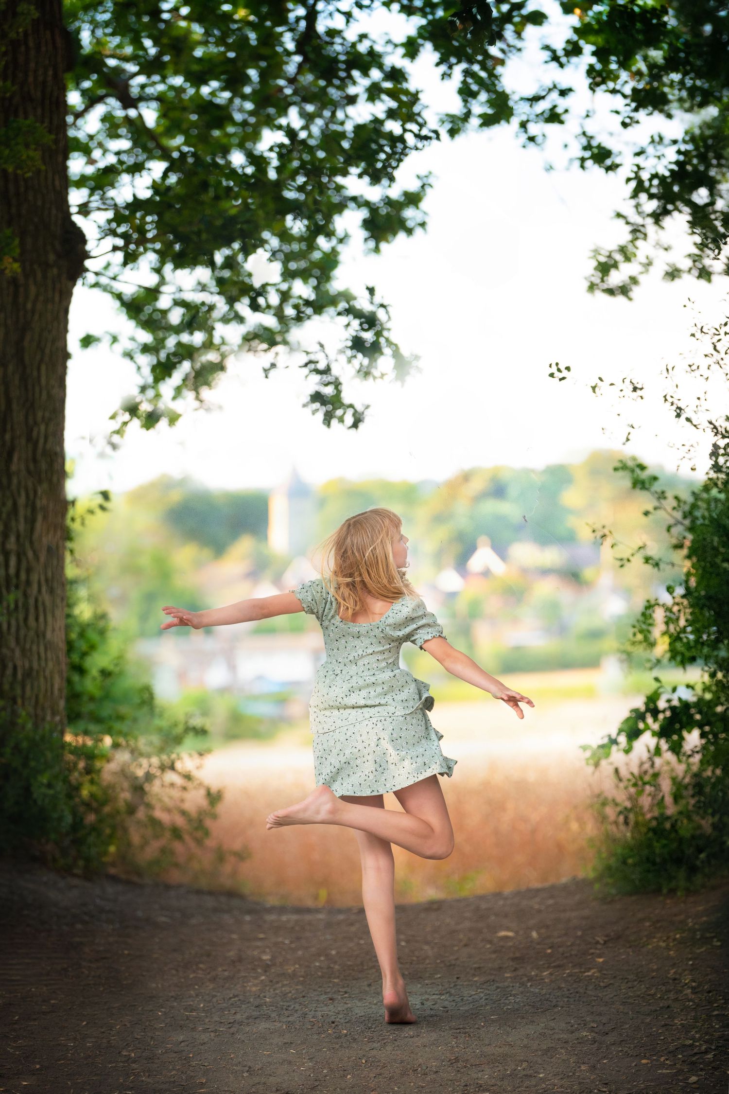 A young woman in a floral dress balances on one leg with arms outstretched on a tree-lined path.