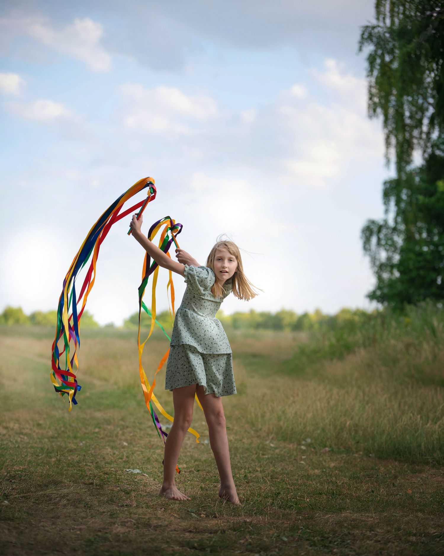 A barefoot girl in a green dress twirls colorful ribbons outdoors in a sunny field.