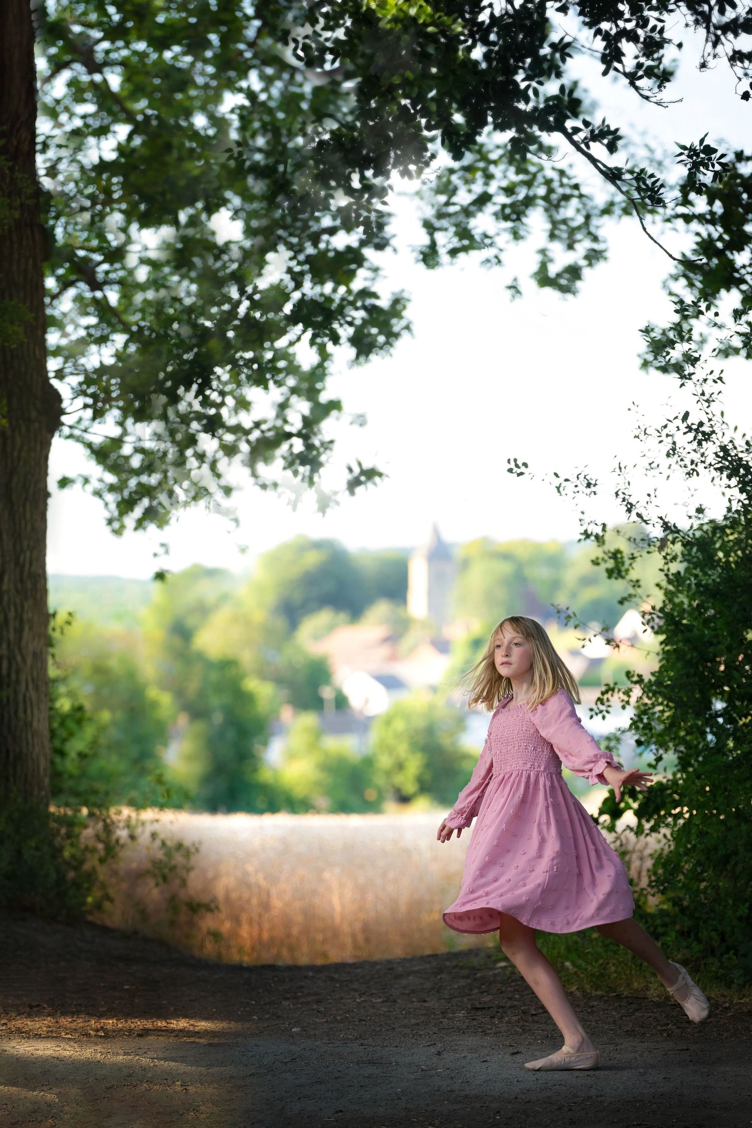 A young girl in a pink dress twirls on a sunlit path framed by trees with a village visible below.