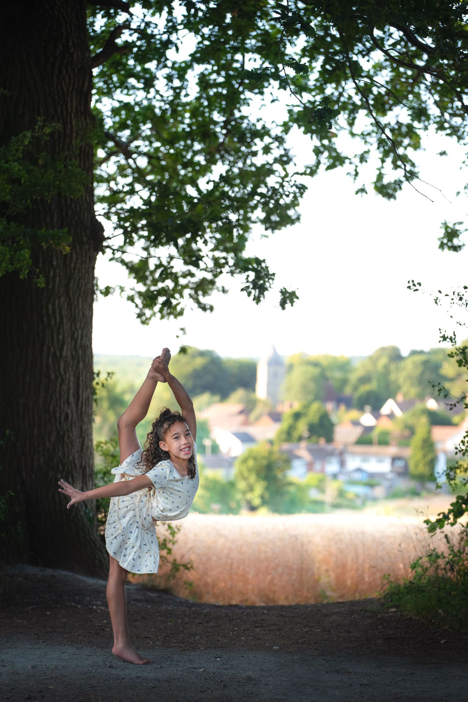Young girl in white dress performs a standing split pose barefoot under a large tree with a village visible in the background.