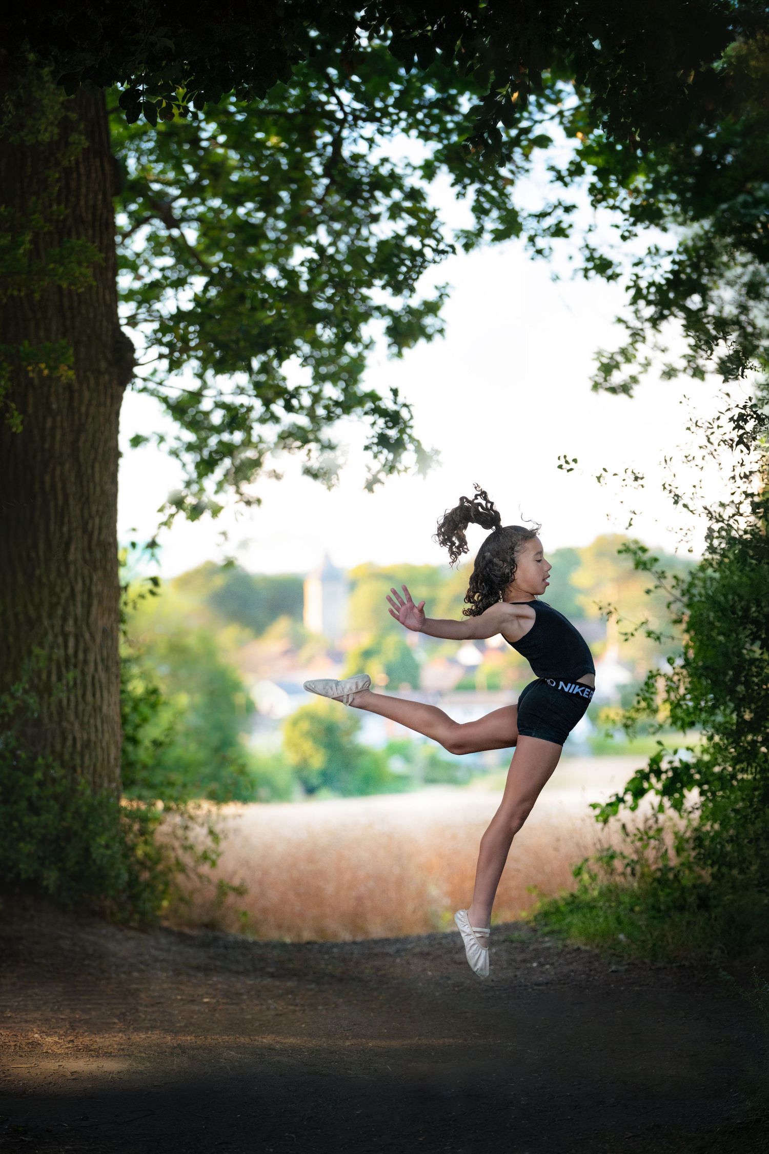 Young girl in black dancewear performs a graceful arabesque leap on a tree-lined path outdoors.