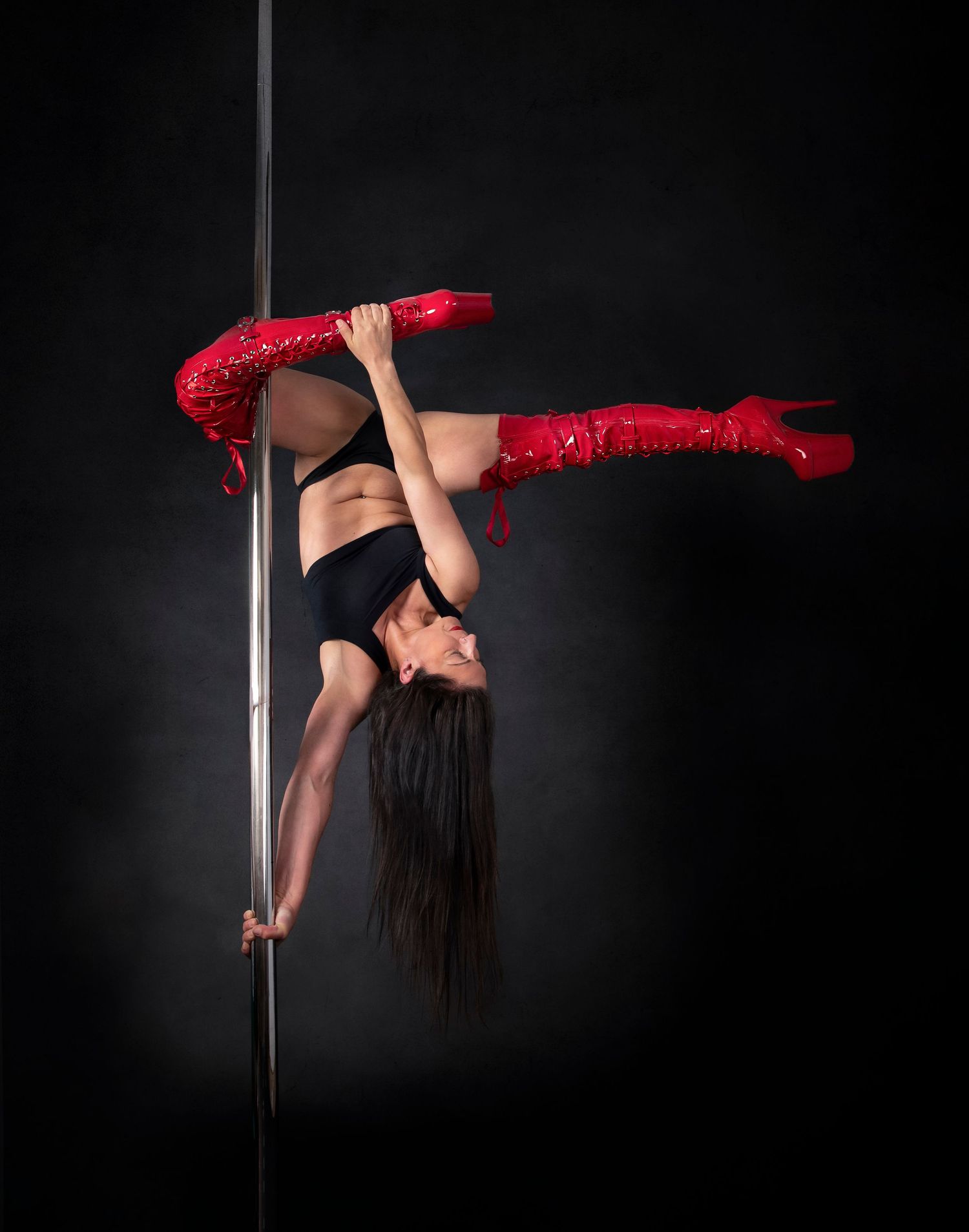 Dancer performs an inverted pose on a pole while wearing red thigh-high boots and black attire against dark background.