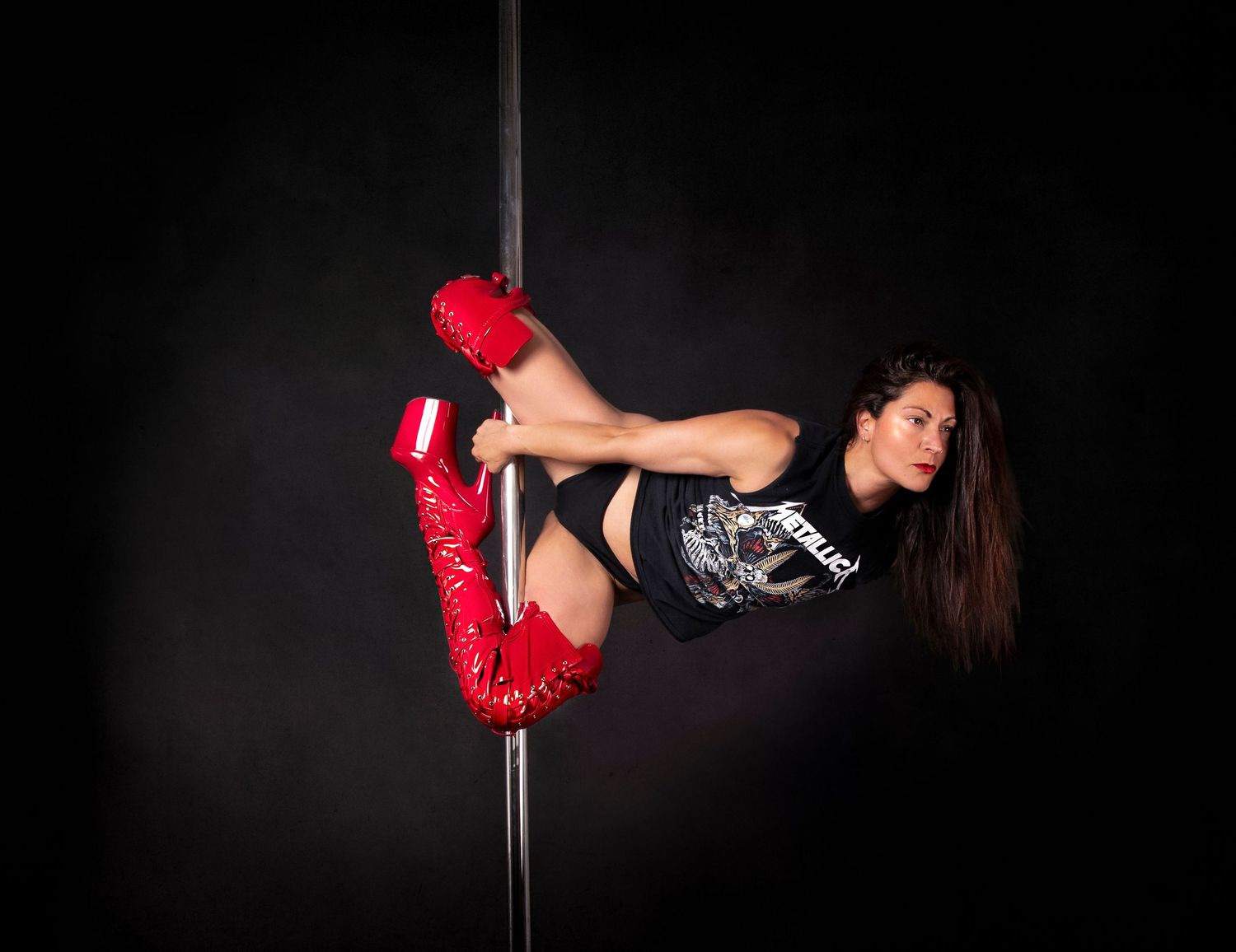 A pole dancer performs an inverted pose wearing red boots and black attire against a dark background.