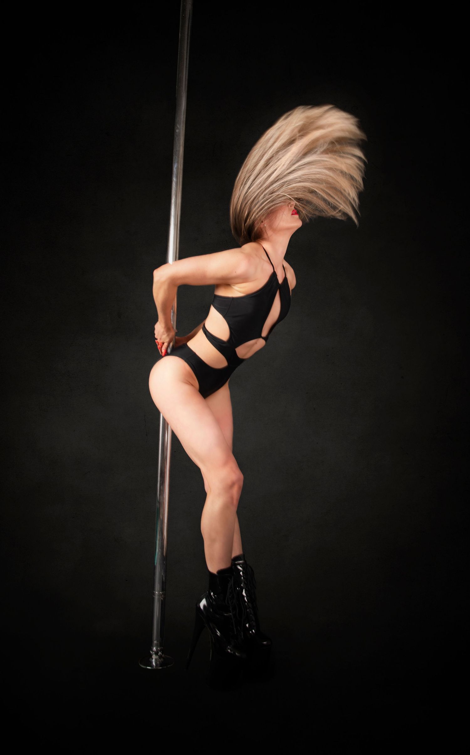 A pole dancer in a black outfit and boots performs a dynamic hair flip move against a dark background.
