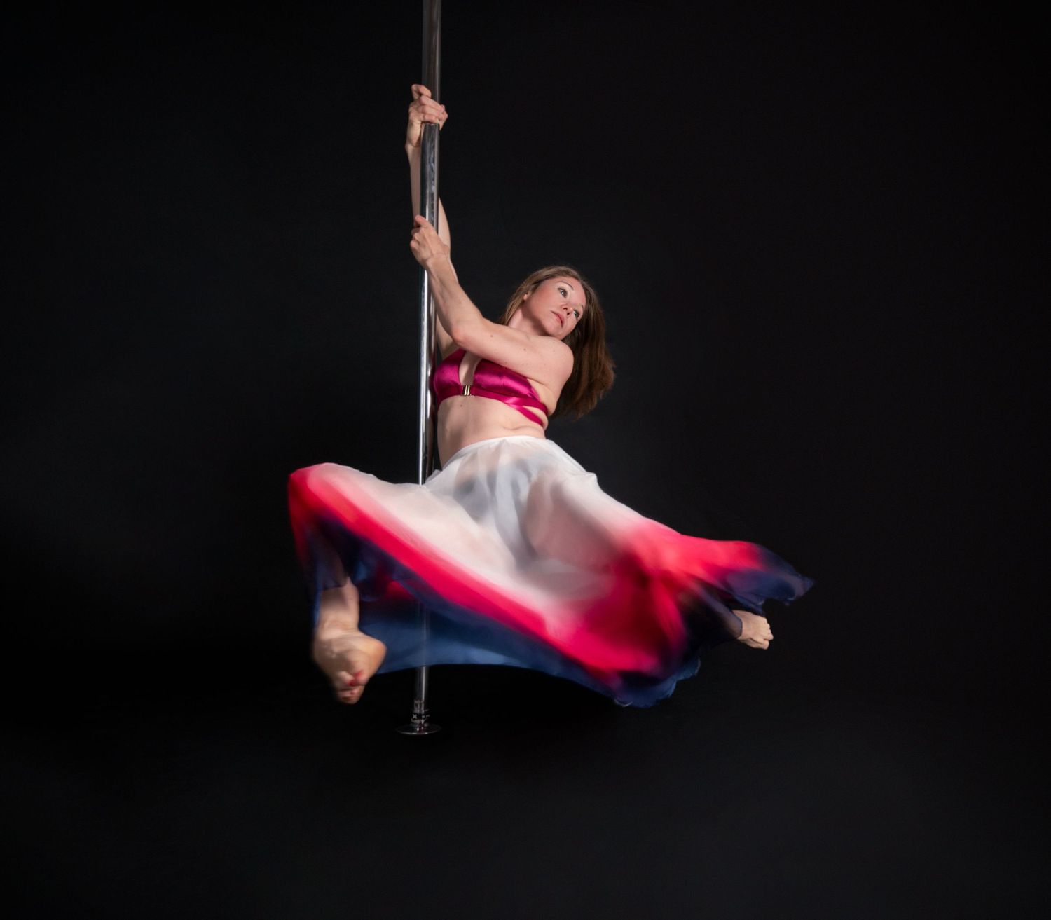 Graceful pole dancer in pink and white dress performs dynamic aerial movement against dark background.