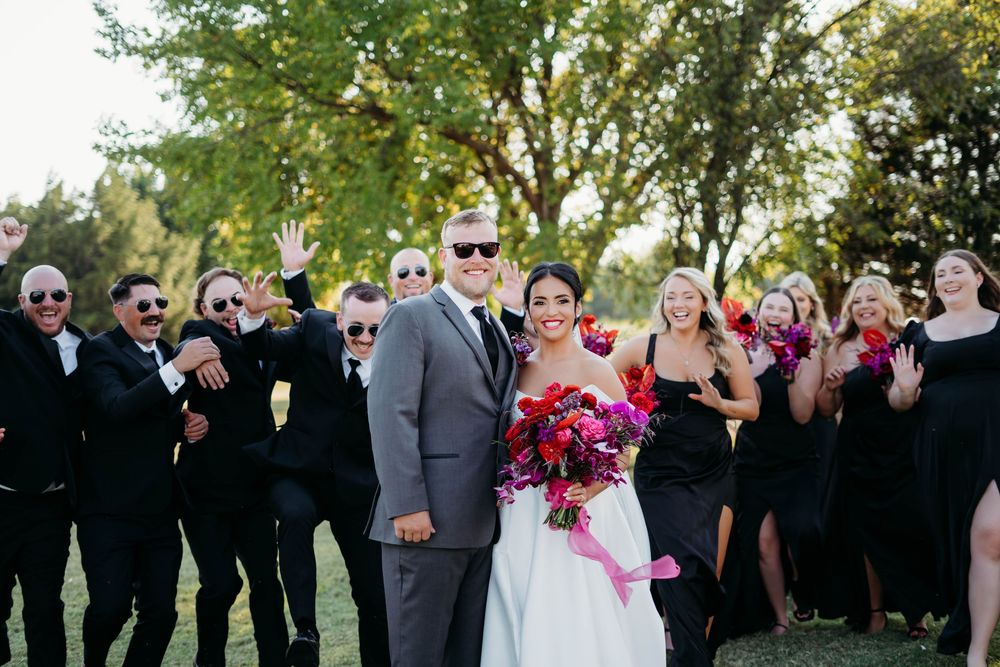 Wedding party celebrates outdoors with bridal couple surrounded by attendants in black dresses and suits.