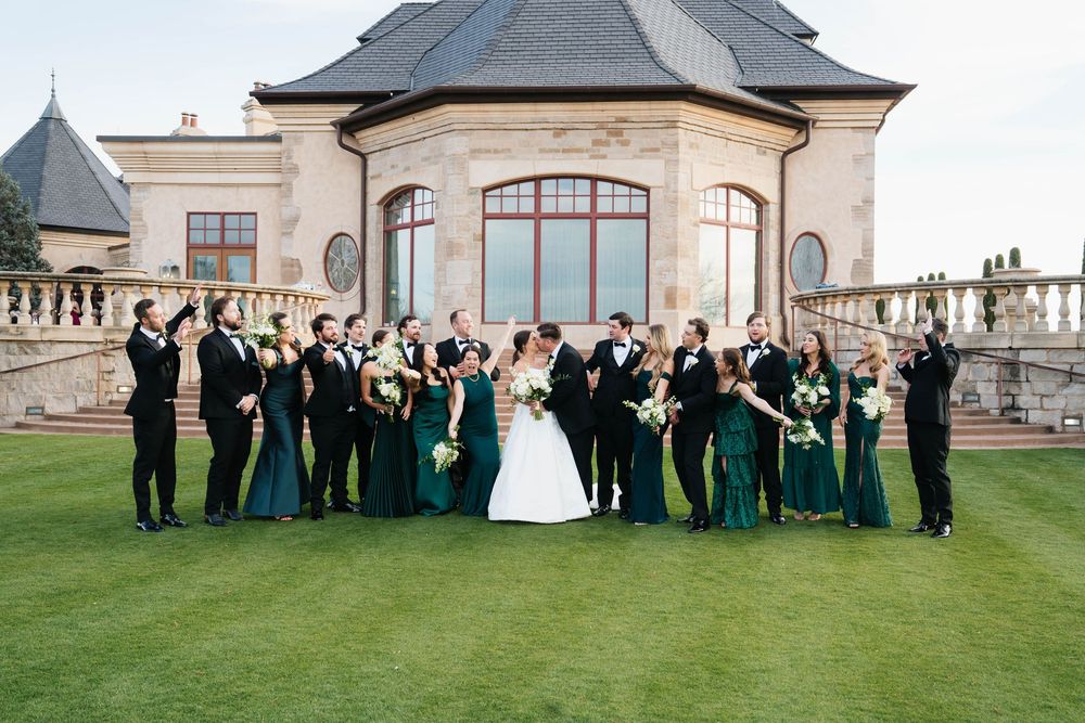Wedding party in emerald green dresses and black tuxedos posing on lawn before elegant stone venue.