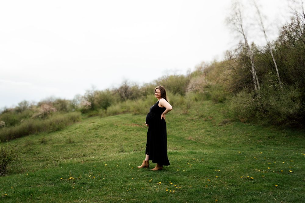 A person in a long black dress stands on a grassy hillside surrounded by spring foliage.