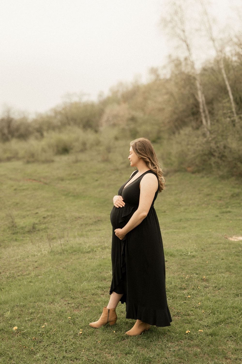 A pregnant woman in a flowing black maxi dress stands in profile on a grassy hillside during golden hour.
