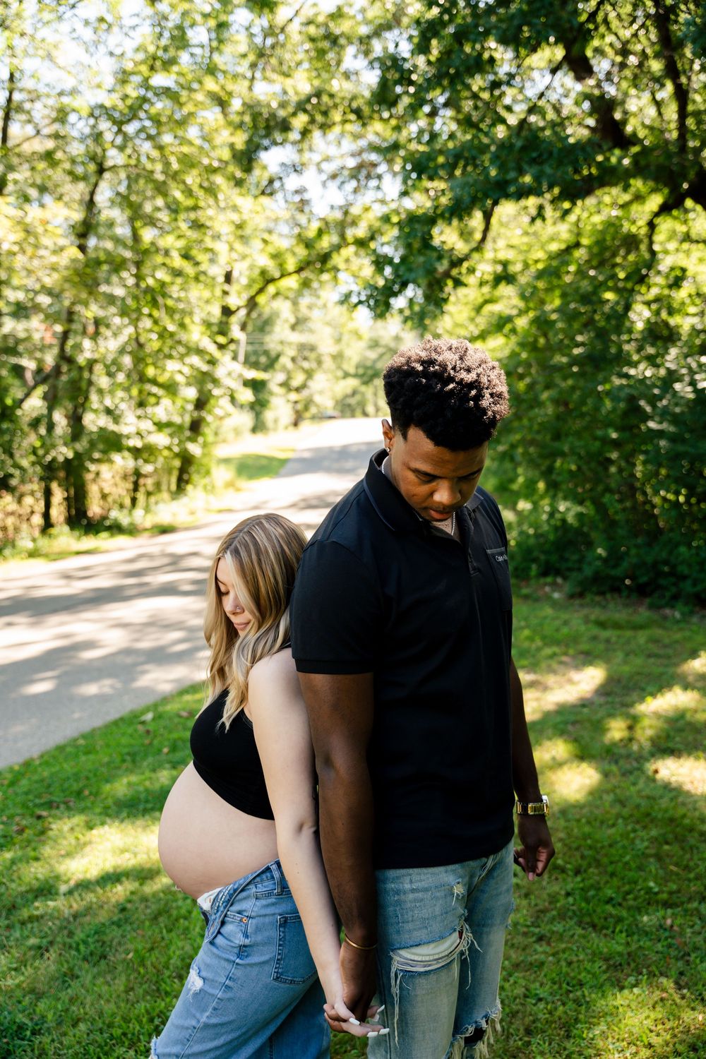 A couple embraces while walking together on a sunlit path through a lush green park on a summer day.