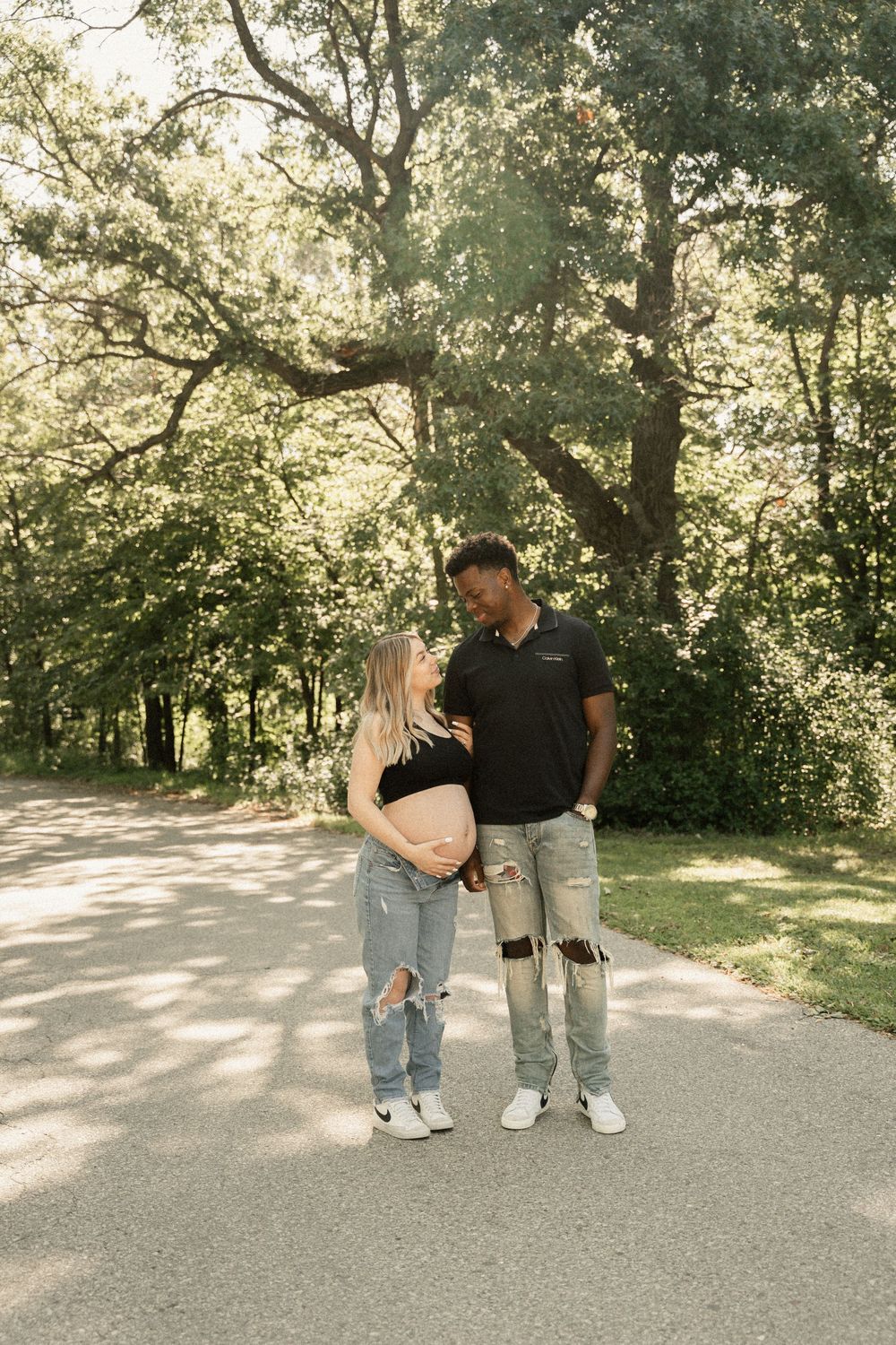 Expectant couple shares a tender moment on a sunlit path surrounded by lush green trees.