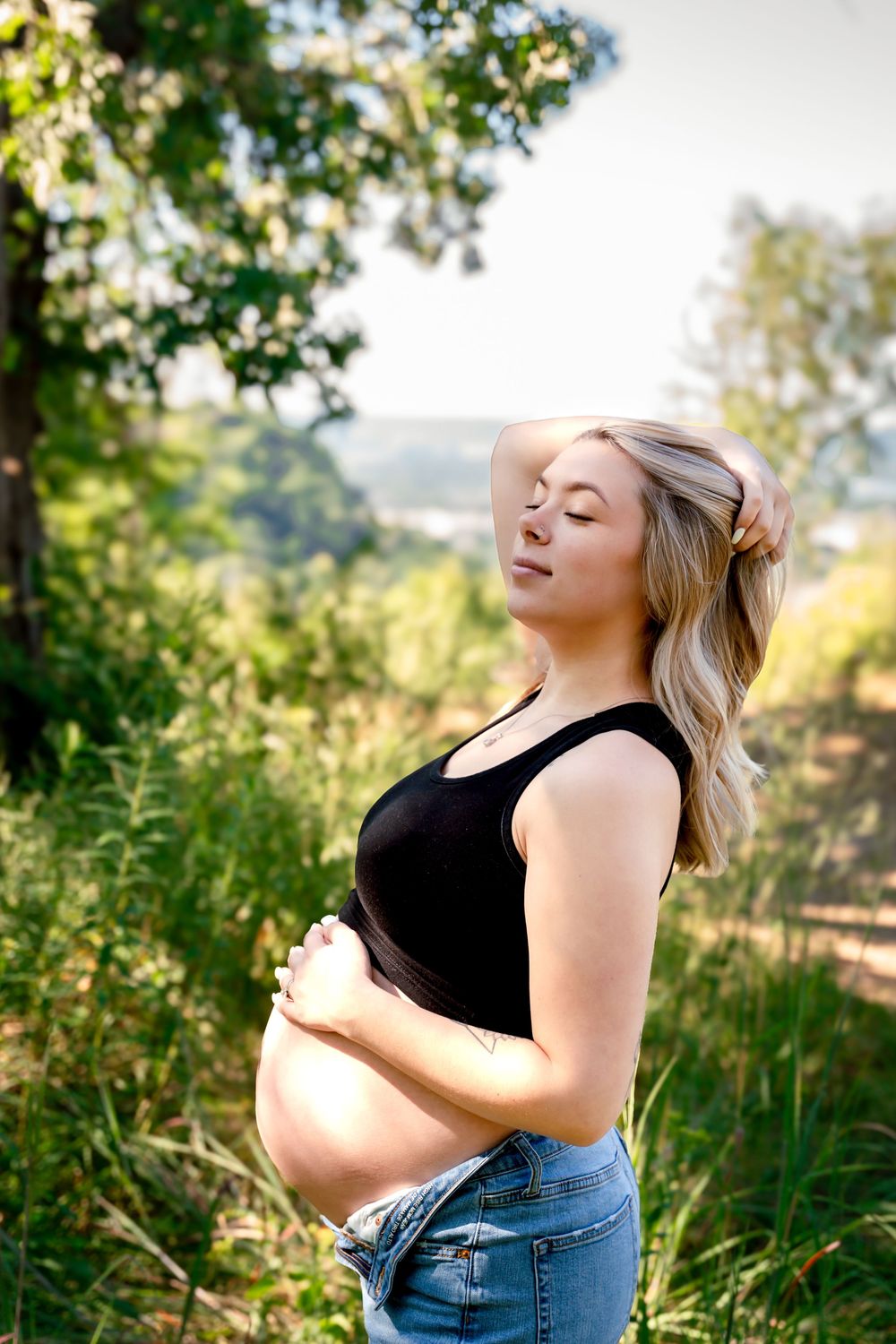 A woman in jeans and black top poses outdoors against a natural backdrop of tall grass and trees.