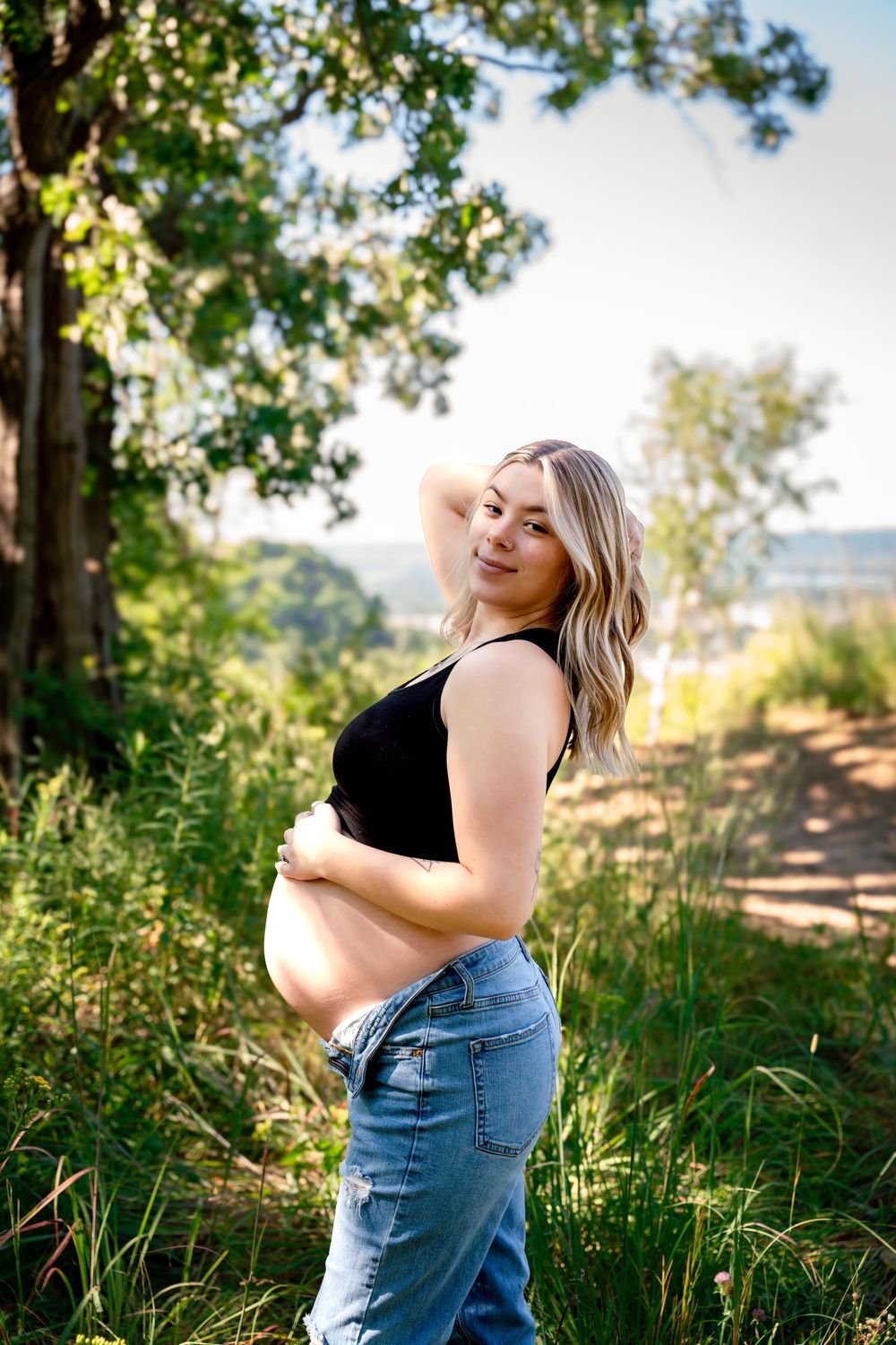 Pregnant woman in denim and black top poses on nature trail with golden sunlight filtering through trees.