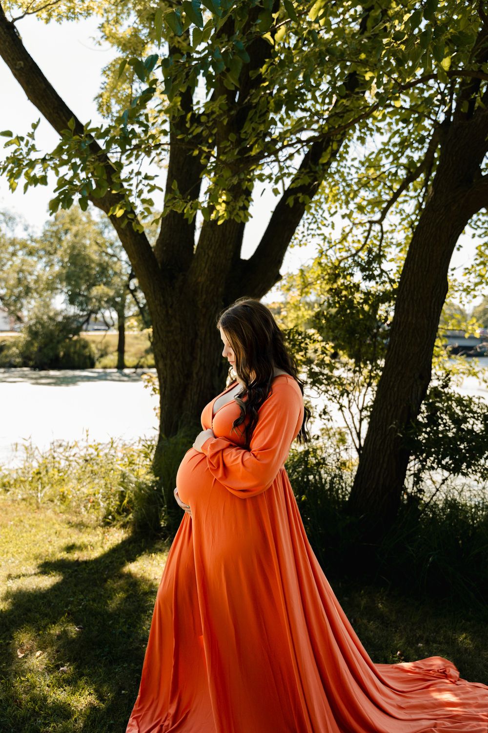 A flowing orange maternity dress catches the sunlight beneath trees in a natural outdoor setting.