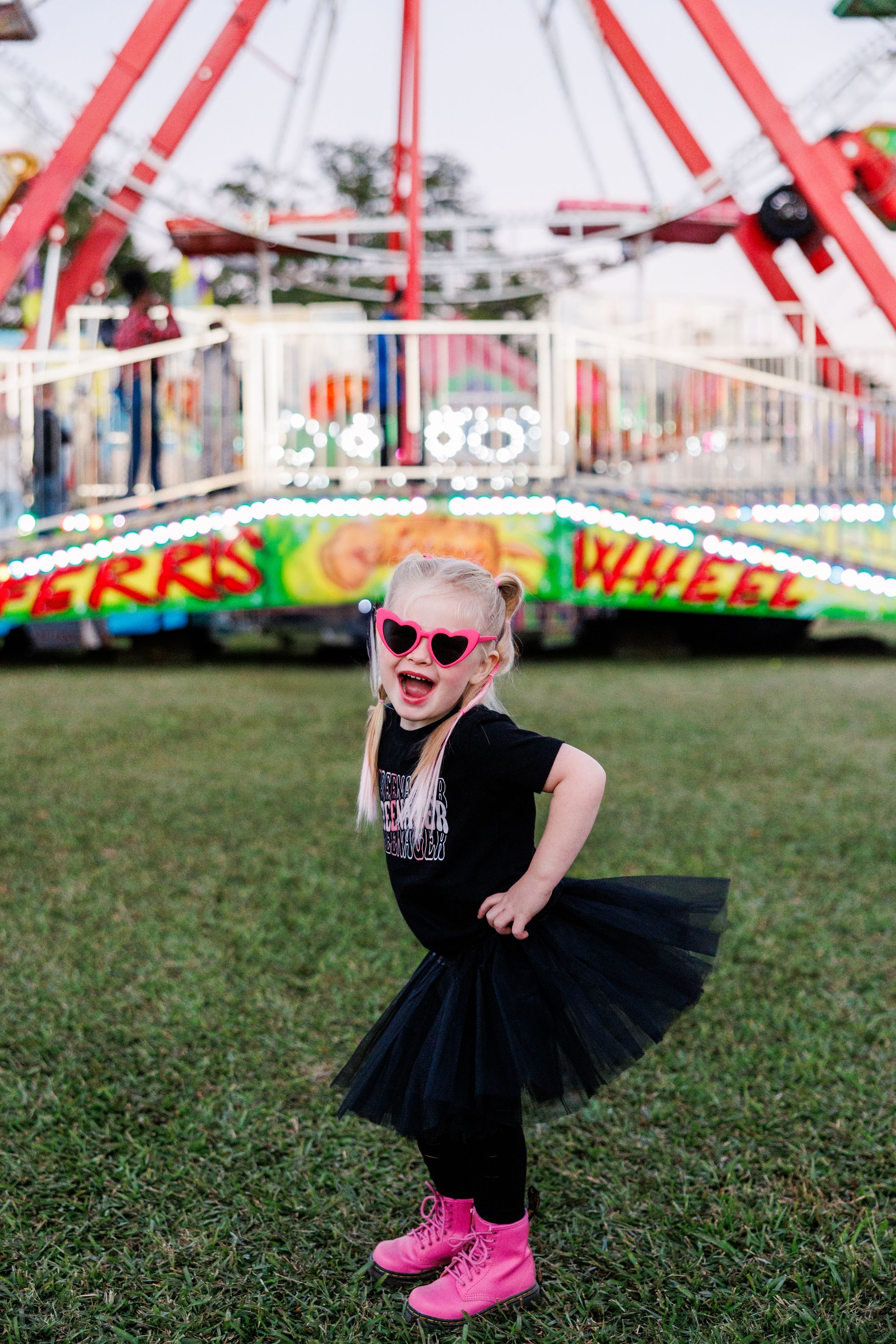 Young dancer in a black tutu and pink boots poses joyfully in front of a carnival ride.