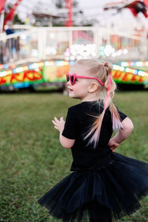 Child in black tutu and pink sunglasses dances at a carnival.