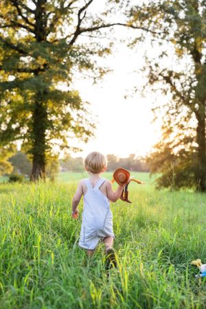 Child in white clothing runs through tall grass at sunset while holding a toy.