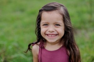 Young child in a burgundy top smiles brightly against a green outdoor background.