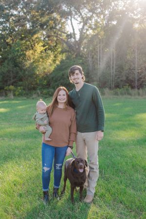 A family stands together with their chocolate lab in a sunlit grassy field.