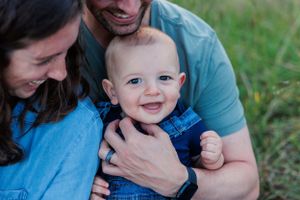 A baby in blue denim overalls smiles joyfully while being held in a loving embrace outdoors.