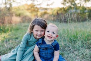 Two young children sit together in grass, sharing a happy moment while wearing casual clothing.