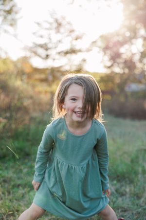 A young child in a green dress plays outdoors with a big smile during golden hour.