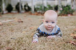 Baby in patterned outfit crawling on dried grass during autumn.