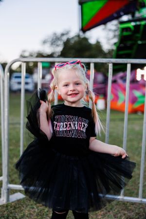 A cheerful child in a black tutu dress poses near carnival barriers.