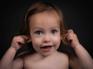 A happy toddler on a black background pulls on their pigtails while smiling playfully at the camera.