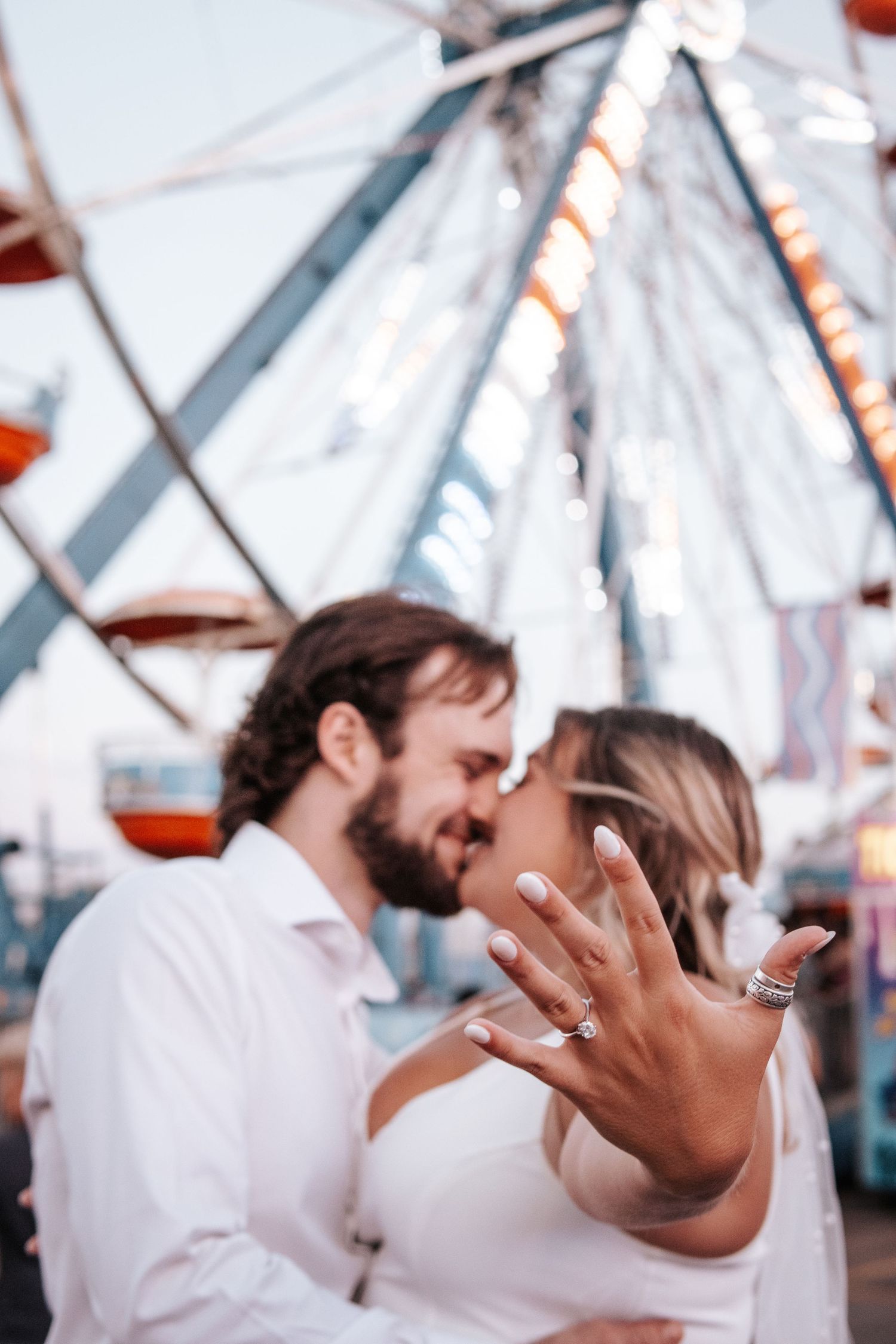 A couple shows off an engagement ring in front of a ferris wheel at twilight.