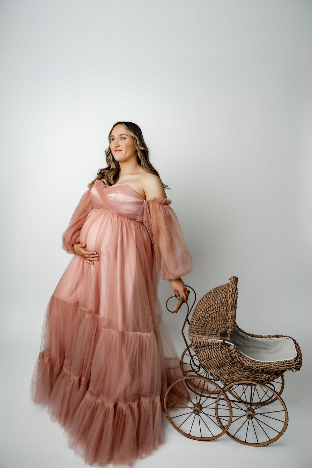 Woman in flowing dusty rose tulle maternity gown poses elegantly next to vintage wicker baby carriage.
