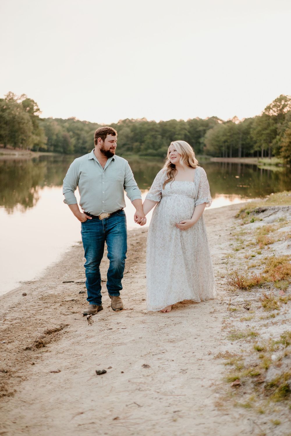 Couple walking hand in hand along lakeside path during sunset maternity photoshoot.