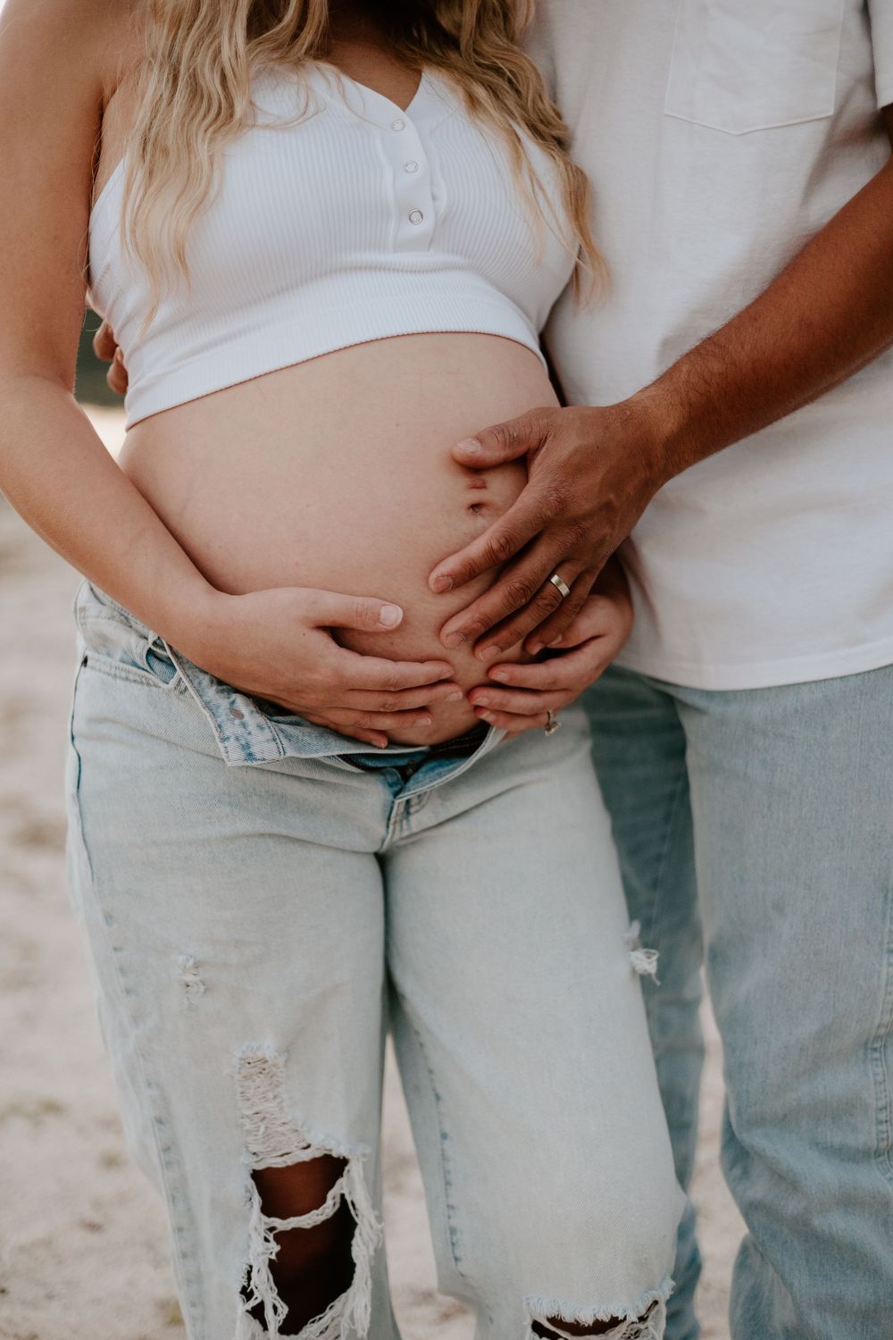Close-up of hands tenderly cradling a pregnant belly while wearing casual ripped jeans and white top.