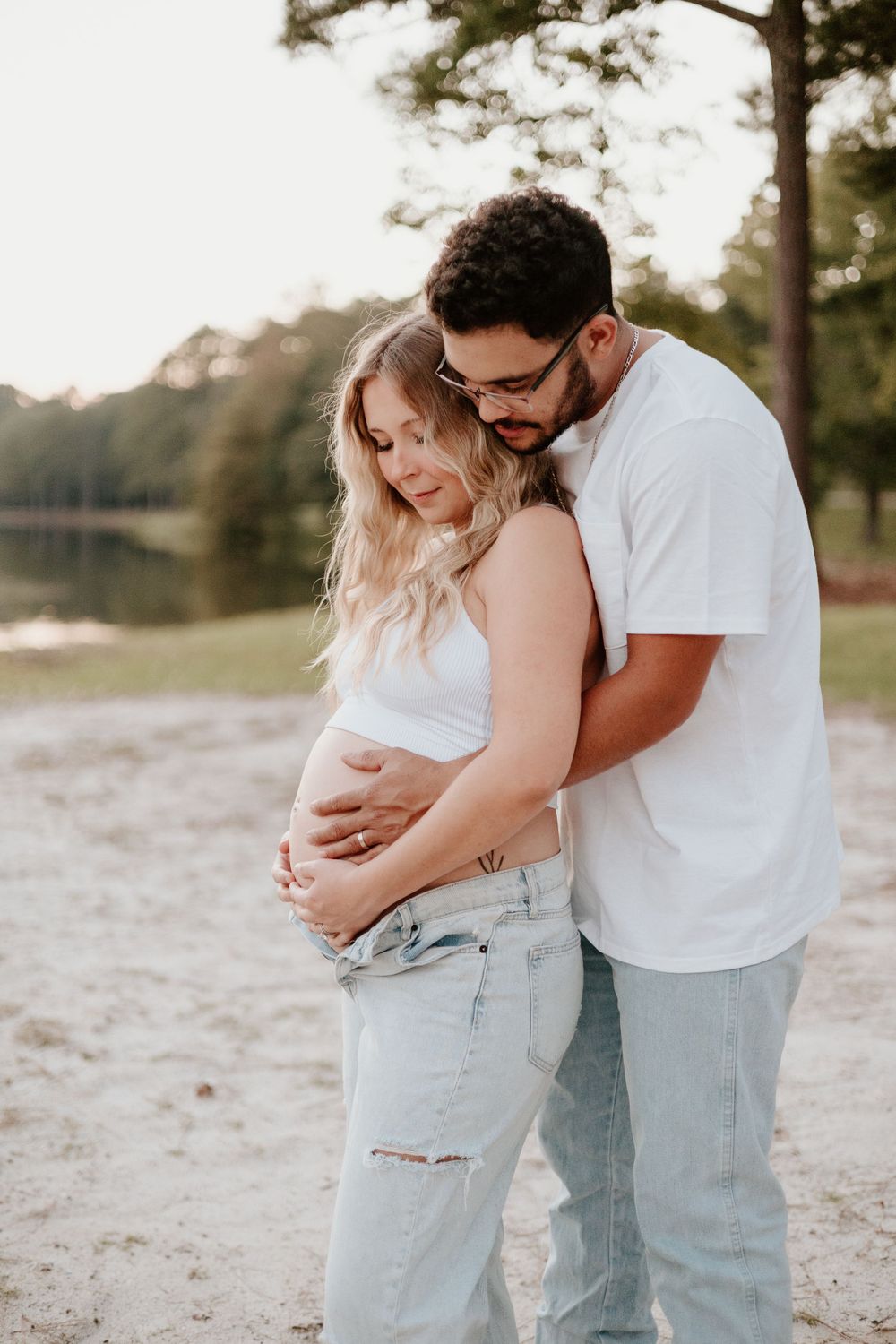 Intimate maternity photo showing couple embracing on beach with gentle water and trees in background.