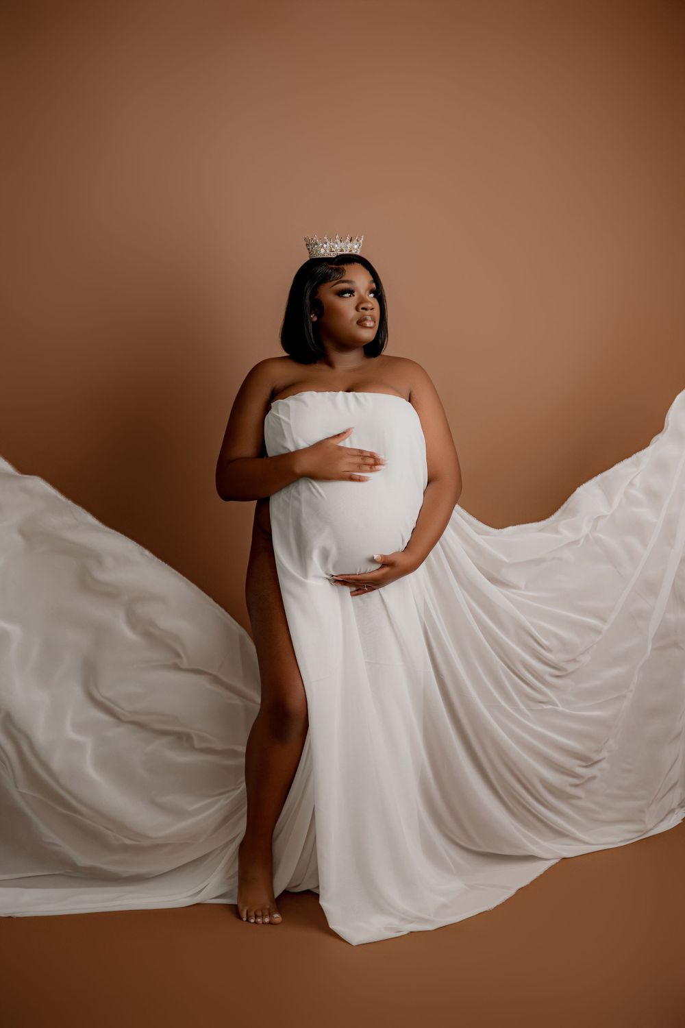 Maternity portrait with flowing white gown and crown against brown backdrop.