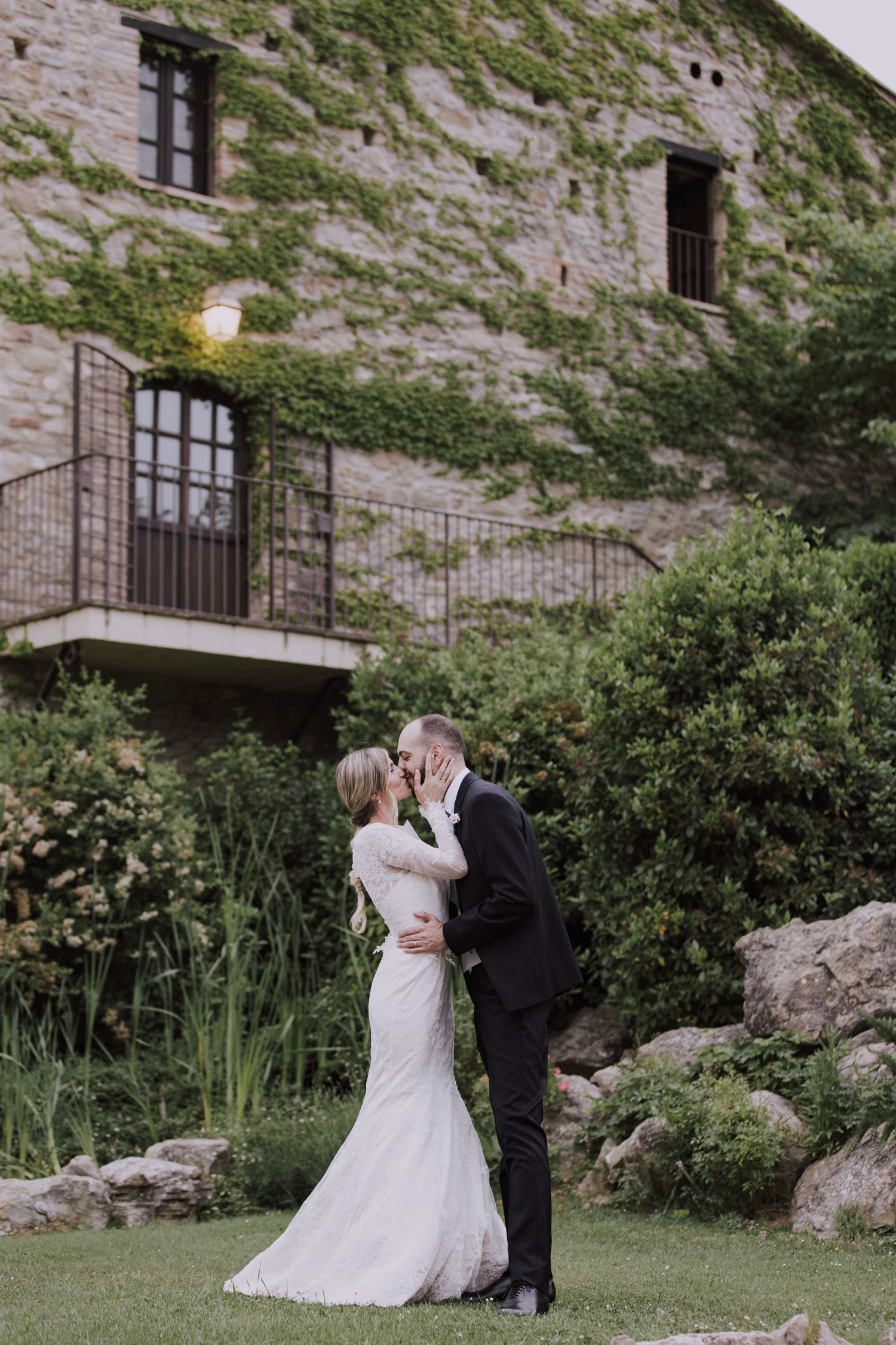 Romantic wedding couple embracing in front of ivy-covered stone building with balcony.