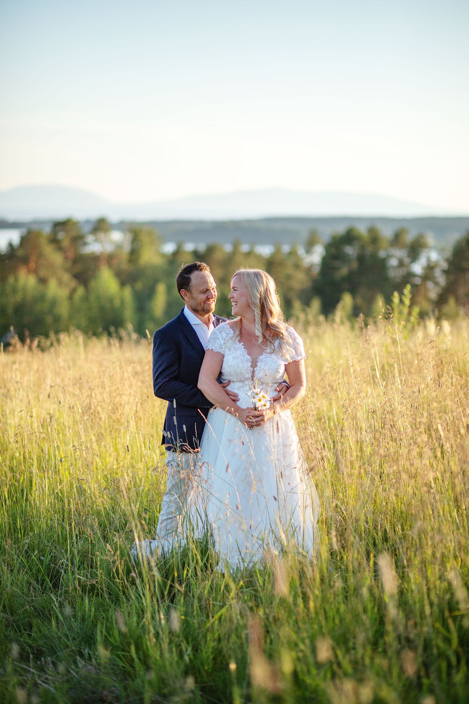 Wedding couple holding heart-shaped balloons on a tree-lined path, smiling at each other in summer greenery.