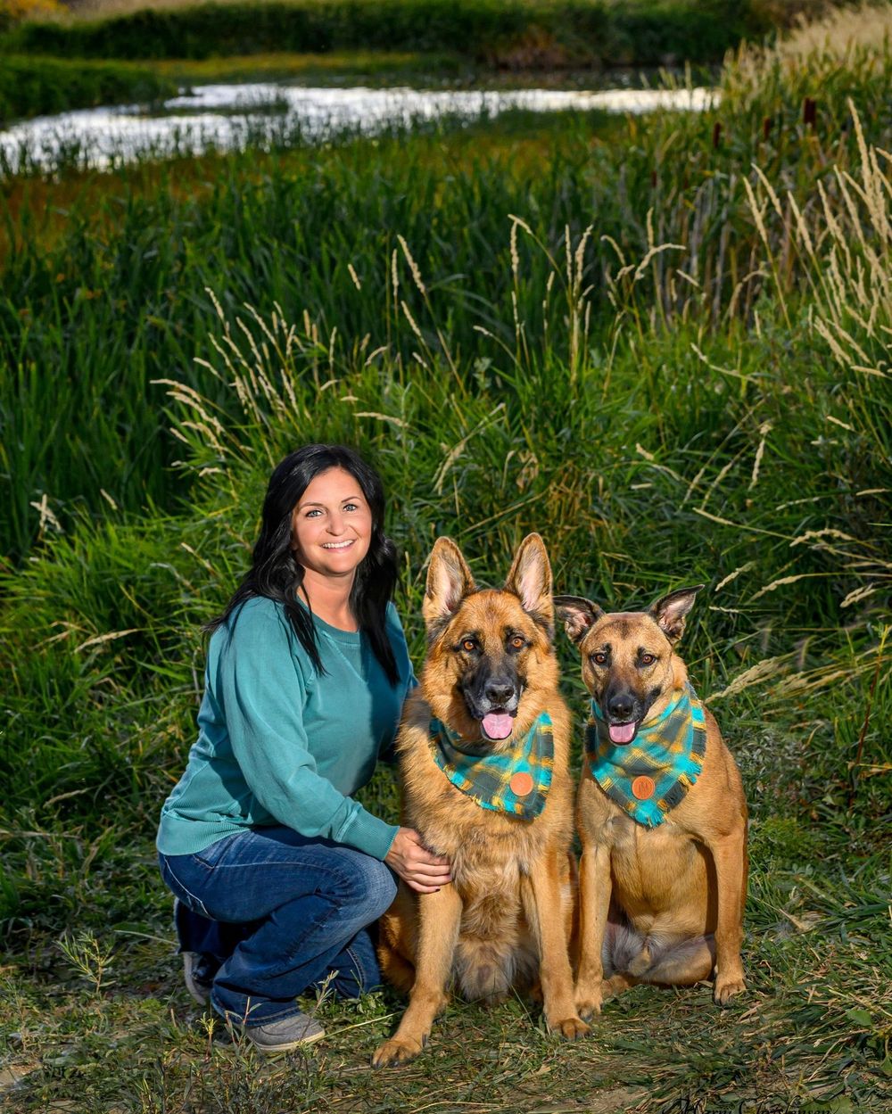 A person sits with two German Shepherd dogs in tall grass near a pond on a sunny day.