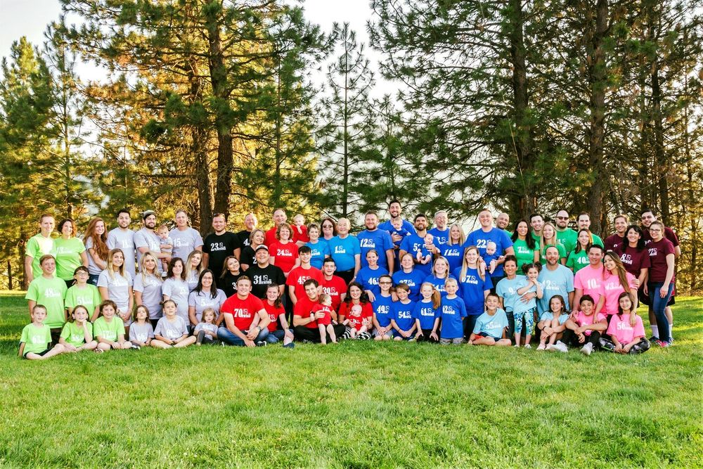 Large group of people wearing colorful t-shirts sit together on a grassy hillside for a group photo.