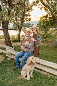 A family sits on a wooden fence with their two dogs at sunset by a lake with trees in the background.
