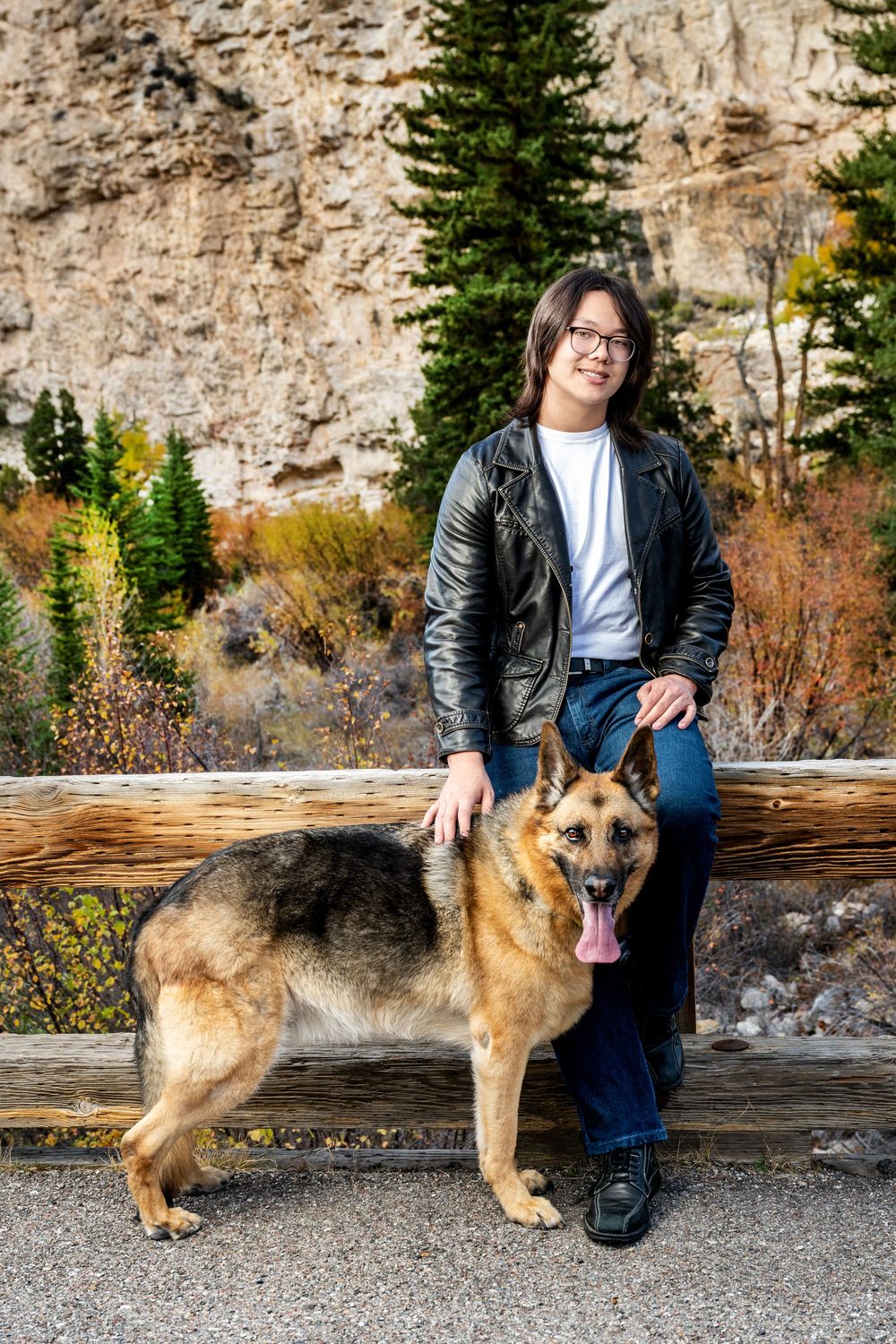 A German Shepherd dog sits with someone on a log bench against a rocky mountain backdrop in autumn scenery.