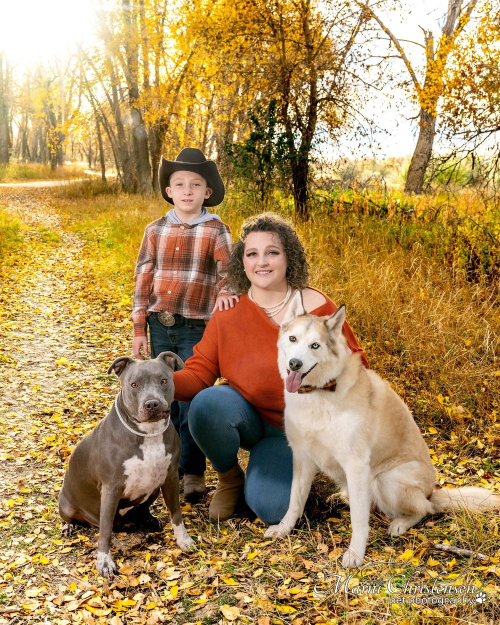 A family and their two dogs sit together on fallen autumn leaves in a golden forest setting.