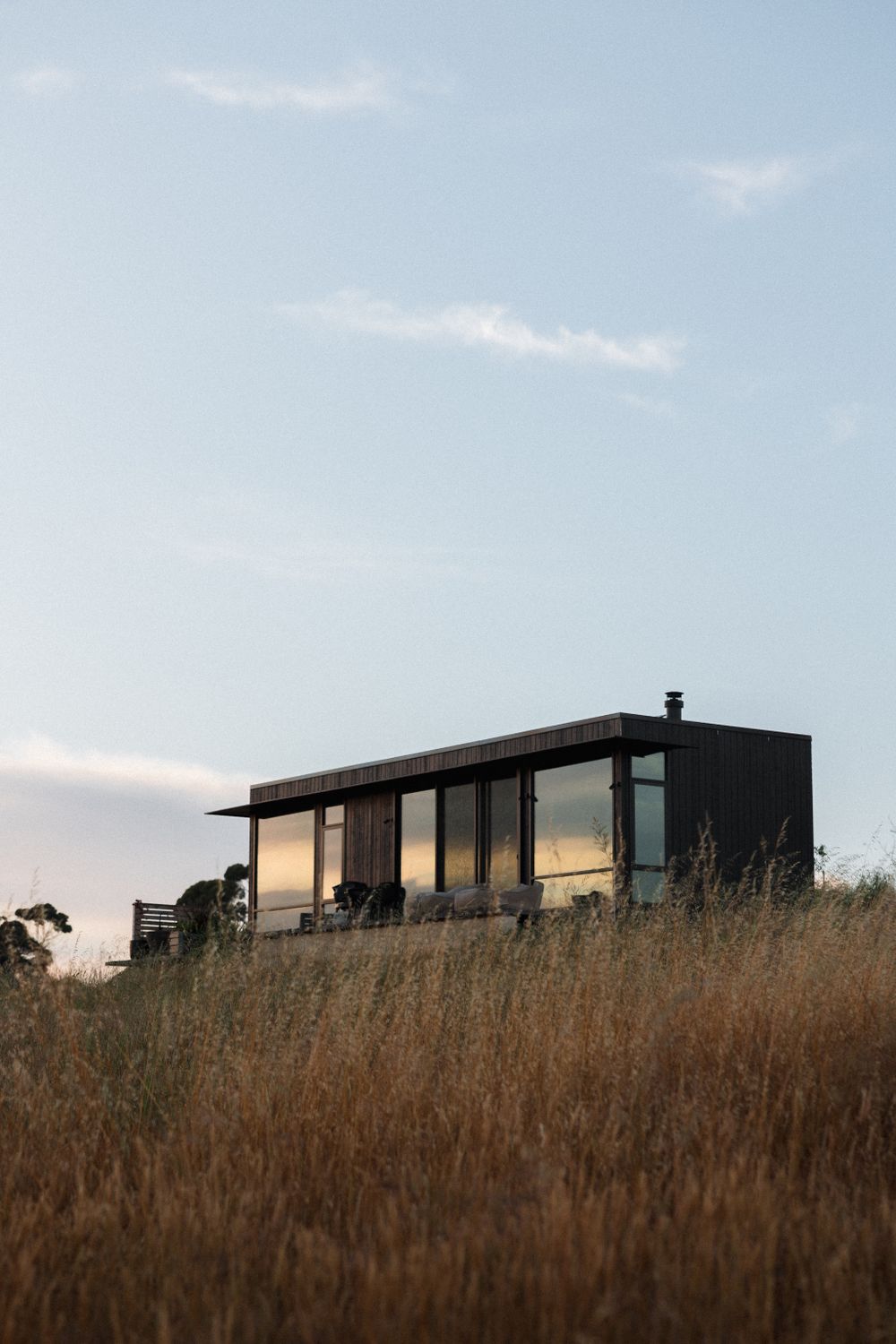 Modern black cabin with large windows perched on a hilltop surrounded by golden grass at sunset.
