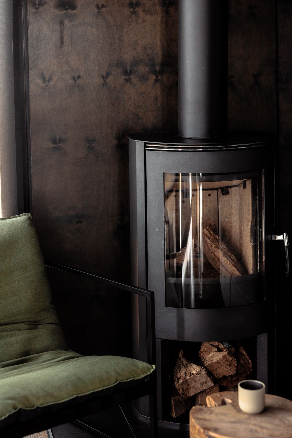 Modern glass wood stove with burning logs next to green velvet armchair.