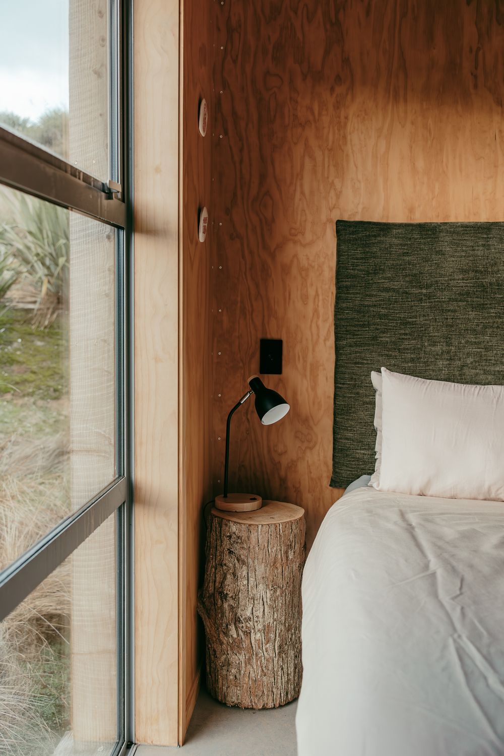 A rustic wood log nightstand sits next to a cozy bed with white linens near a window.