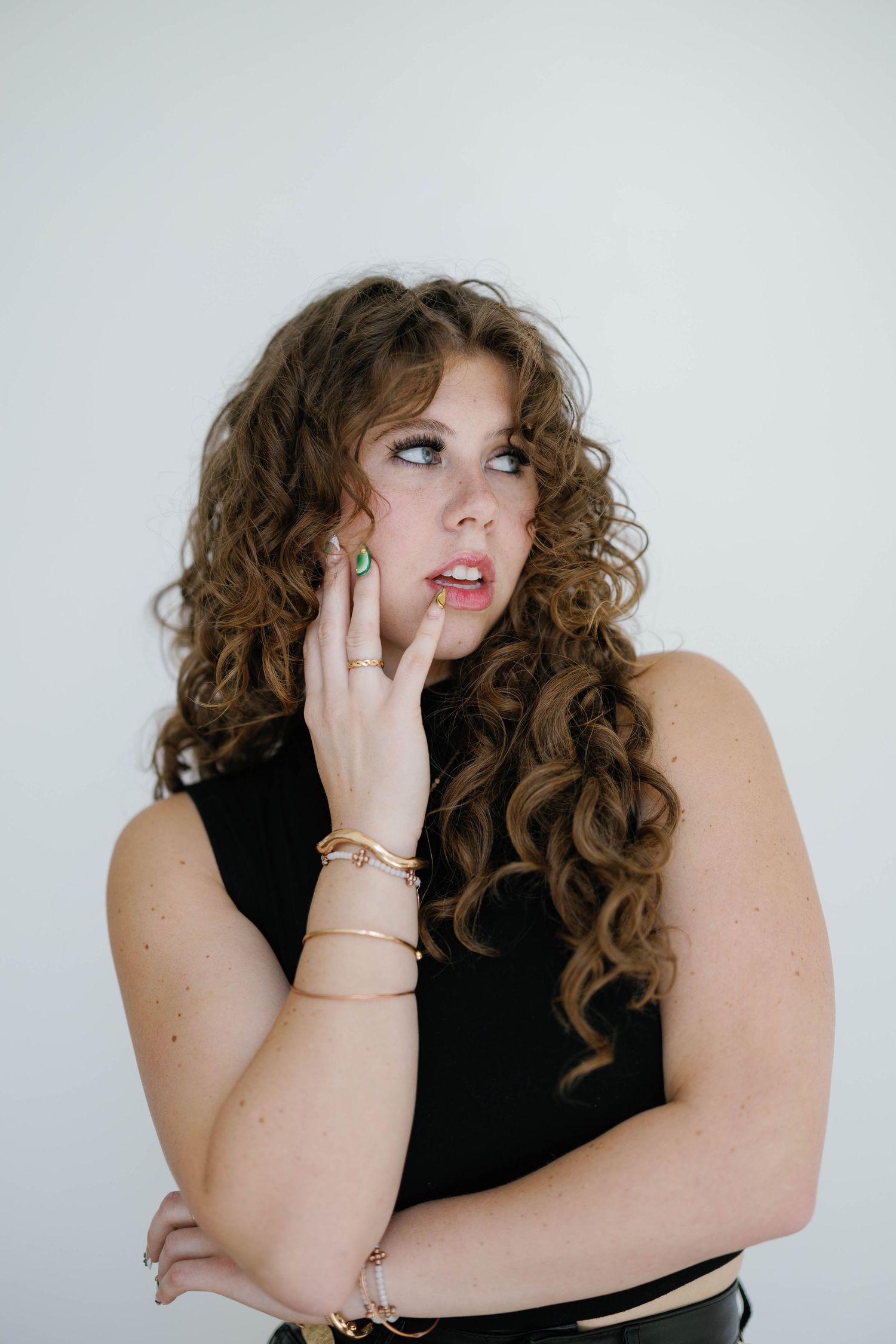 Young woman with curly auburn hair poses thoughtfully in black sleeveless top with gold bracelets and green ring.