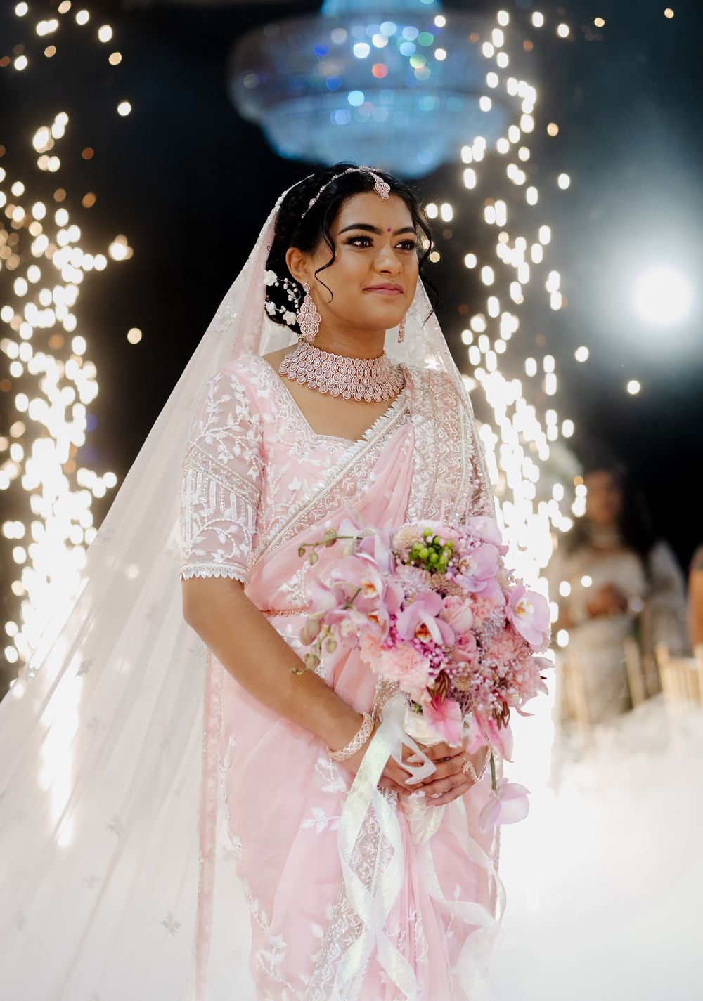 A bride in a pink and white embellished sari holds a pink bouquet under sparkling lights at an elegant wedding celebration.