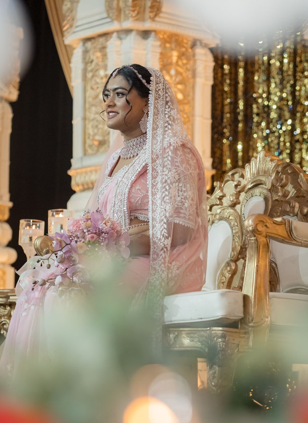 Traditional wedding ceremony with couple seated in ornate mandap decorated with pink flowers and golden details.