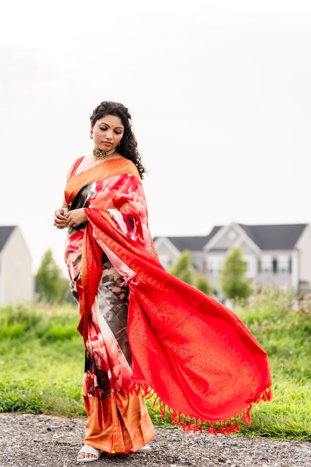 A person wearing a flowing red and orange sari poses gracefully outdoors with houses in the background.