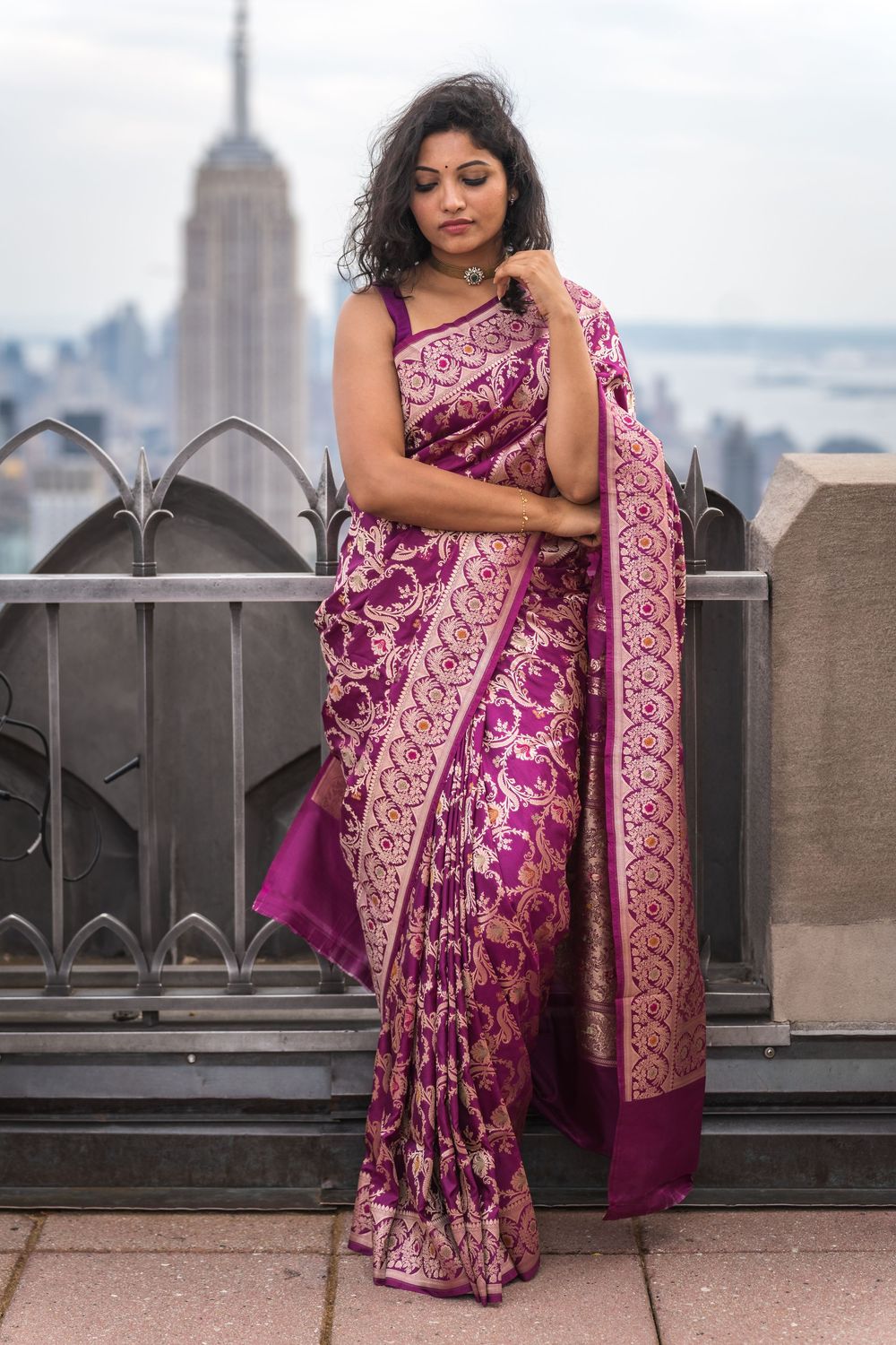A woman in a purple and gold embroidered sari poses on a rooftop with the New York City skyline in the background.